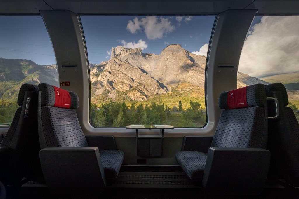 Vue depuis l'intérieur d'une voiture de train première classe : sièges face à face, hublot panoramique sur des montagnes ensoleillées et une forêt.